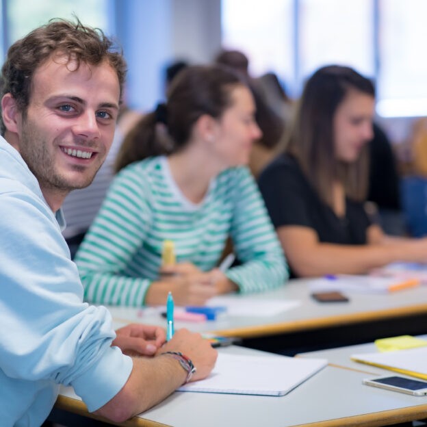Un étudiant vêtu d'un pull bleu, souriant, est en avant-plan, en train d'écrire sur une feuille de papier. Derrière lui, deux jeunes étudiantes sourient également.