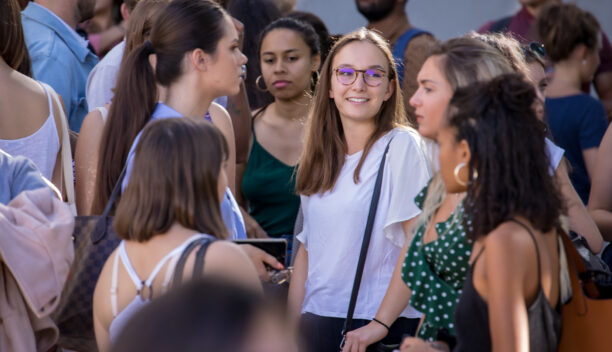 Un groupe d'étudiants est debout, majoritairement des filles. Une fille, au milieu, portant un t-shirt blanc et un sac noir, ainsi que des lunettes, sourit. Image pour le parcours d'engagement citoyen étudiant.