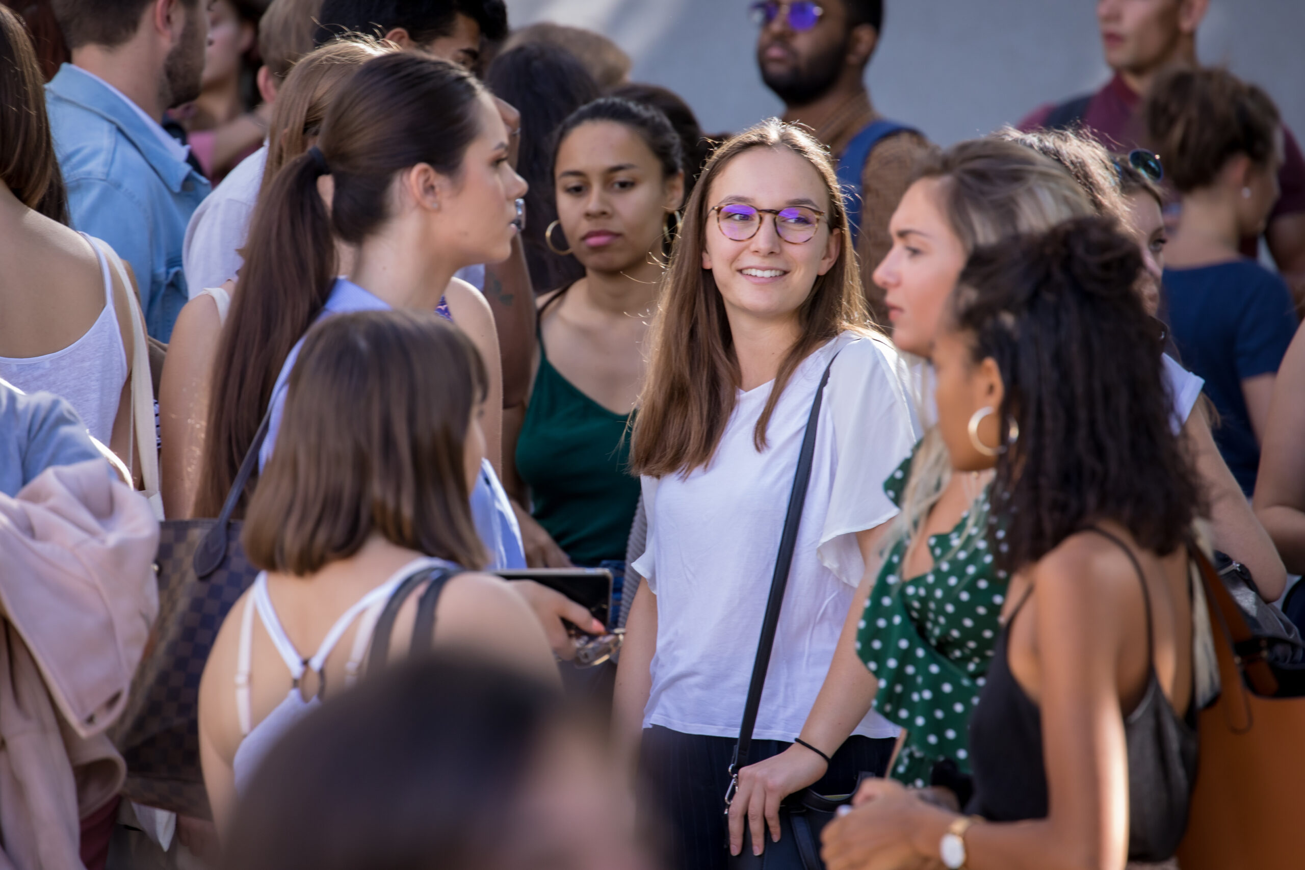 Un groupe d'étudiants est debout, majoritairement des filles. Une fille, au milieu, portant un t-shirt blanc et un sac noir, ainsi que des lunettes, sourit. Image pour le parcours d'engagement citoyen étudiant.