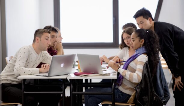 Des jeunes étudiants autour d'une table en train de travailler. Au premier plan, une jeune étudiante aux cheveux bouclés est devant son ordinateur, tandis qu'un jeune étudiant en pull blanc est également devant son ordinateur. Un des étudiants est debout et semble écouter une explication de la jeune étudiante aux cheveux bouclés.